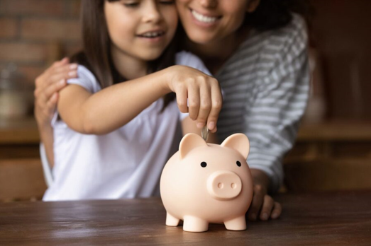 Caucasian mother and daughter with brown hair placing a coin inside of a pink piggy bank.